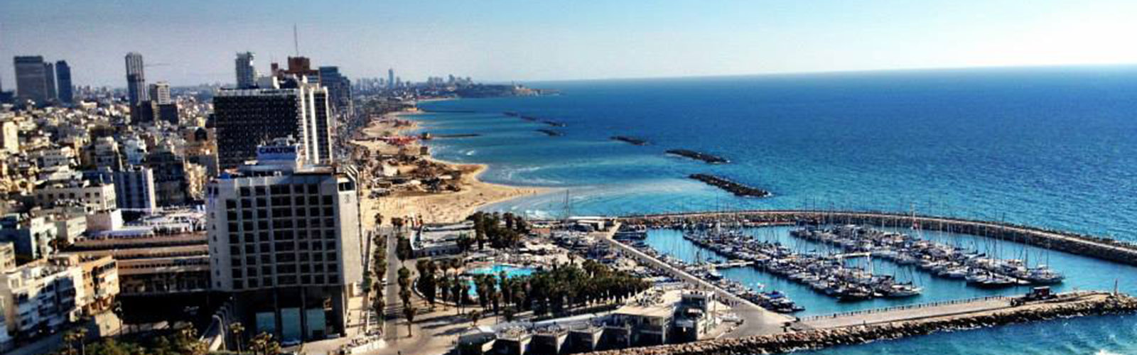 A costal city, with tall buildings on the left side, a sandy beach in the middle, and a crowded harbor leading into a sparkling blue ocean. 